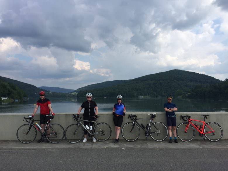 Czworo rowerzystów z rowerami szosowymi na tle jeziora i zalesionych wzgórz